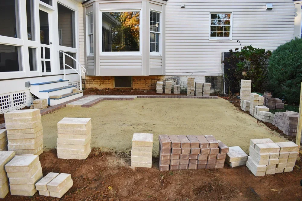 Residential patio foundation prep with organized concrete pavers and retaining wall blocks staged on prepared ground