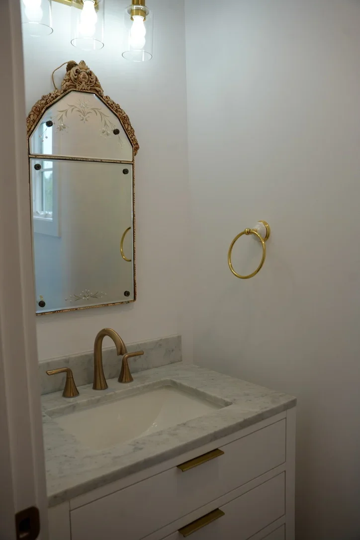 Renovated bathroom vanity with marble countertop, white cabinetry, and ornate brass-framed mirror